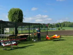 Drei Modellflugzeuge auf einer Rasenfläche, daneben ein älterer Mann mit Hut, im Hintergrund Zaun und Bäume