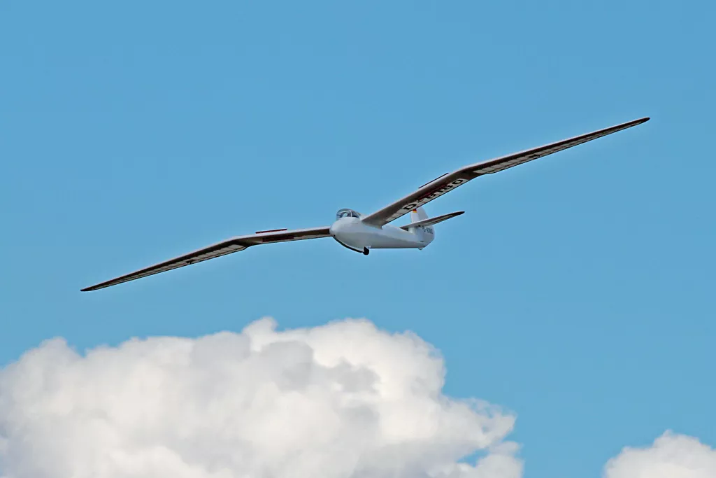 Wei&szlig;es Segelflugzeug mit langen Tragfl&auml;chen fliegt vor blauem Himmel und einer gro&szlig;en wei&szlig;en Wolke