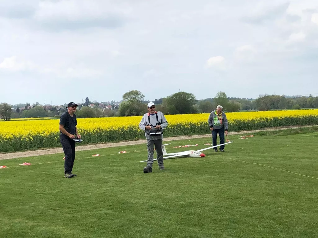 Drei Modellflieger mit Segelflugzeug auf grüner Wiese, gelbes Rapsfeld im Hintergrund, Sommertag