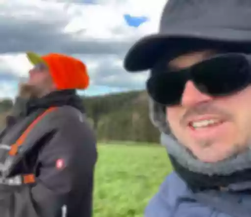 Zwei Personen in Winterkleidung auf Wiese, Wald im Hintergrund. Einer mit oranger Mütze, einer mit schwarzer Sonnenbrille und Fellkragen.