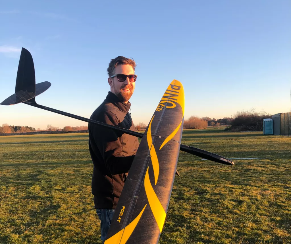 Modellflug-Enthusiast mit gro&szlig;em grau-gelben Segelflugmodell auf Vereinsgel&auml;nde bei Abendd&auml;mmerung