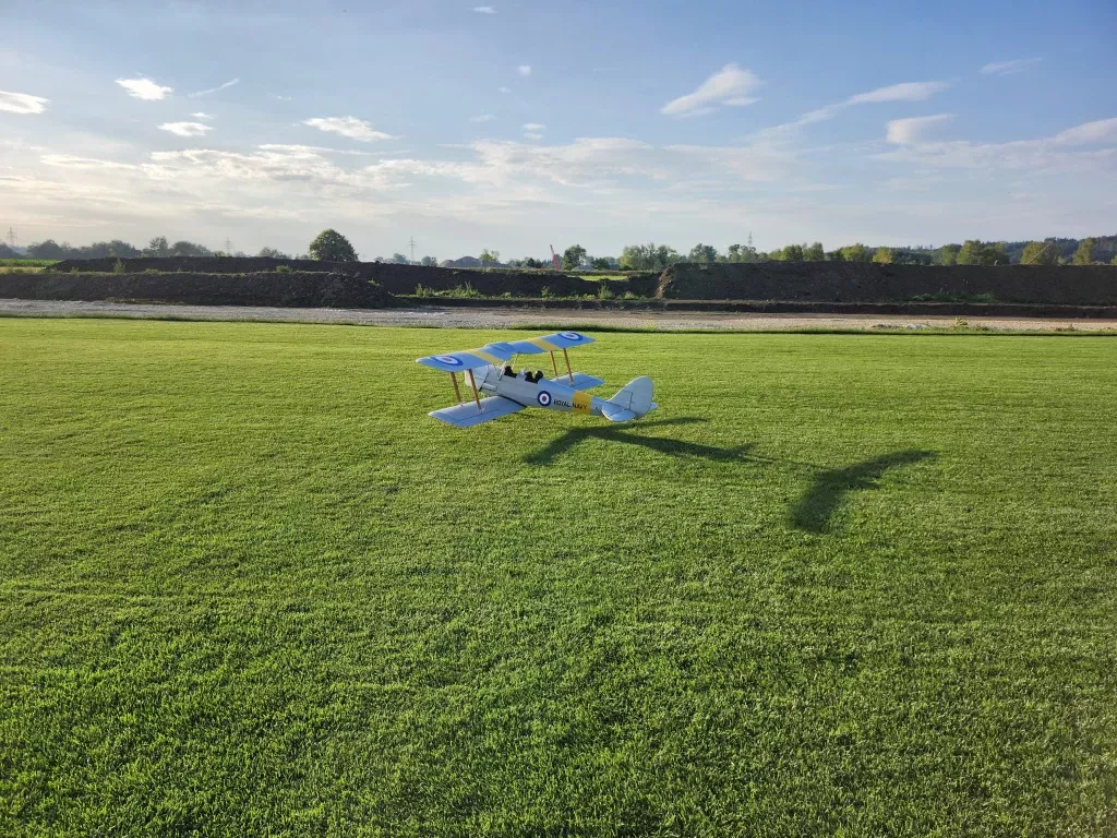Blau-wei&szlig;es Modellflugzeug im Stil eines historischen Doppeldeckers auf einer gr&uuml;nen Rasenfl&auml;che, mit Schatten und Landschaft im Hintergrund