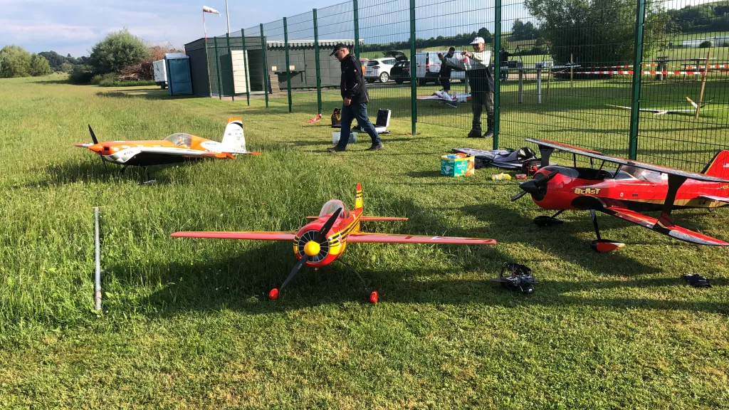 Mehrere bunte Modellflugzeuge auf einer gr&uuml;nen Rasenfl&auml;che, eingez&auml;unt mit Metallzaun, unterschiedliche Gr&ouml;&szlig;en und Designs