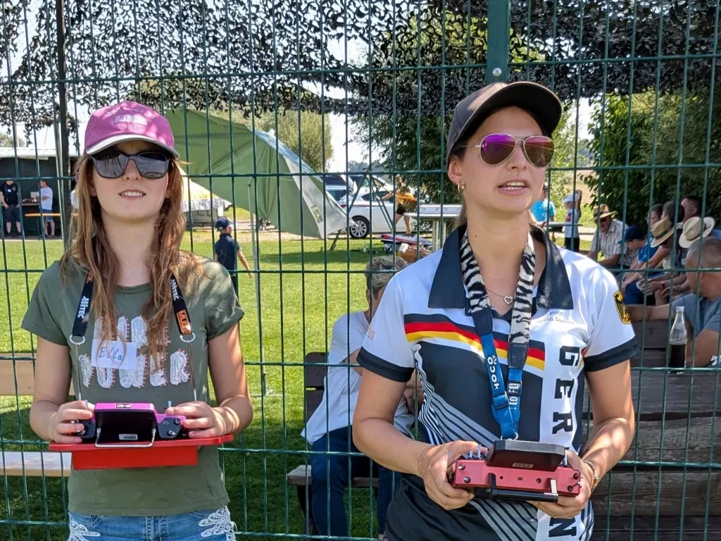 Zwei Frauen mit Sonnenbrillen und Caps stehen hinter Zaun, halten rote Fernsteuerungen f&uuml;r Modellflugzeuge