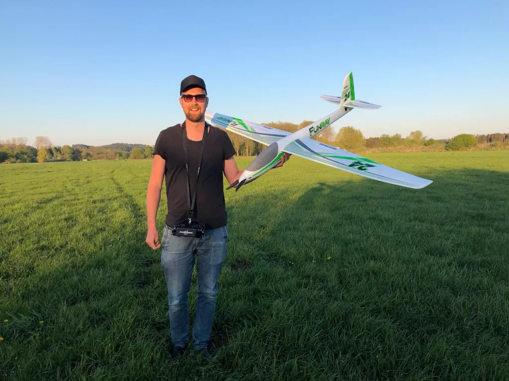 Person mit schwarzem T-Shirt und Basecap h&auml;lt ein gr&uuml;n-wei&szlig;es Modellflugzeug auf einer gr&uuml;nen Wiese mit B&auml;umen im Hintergrund