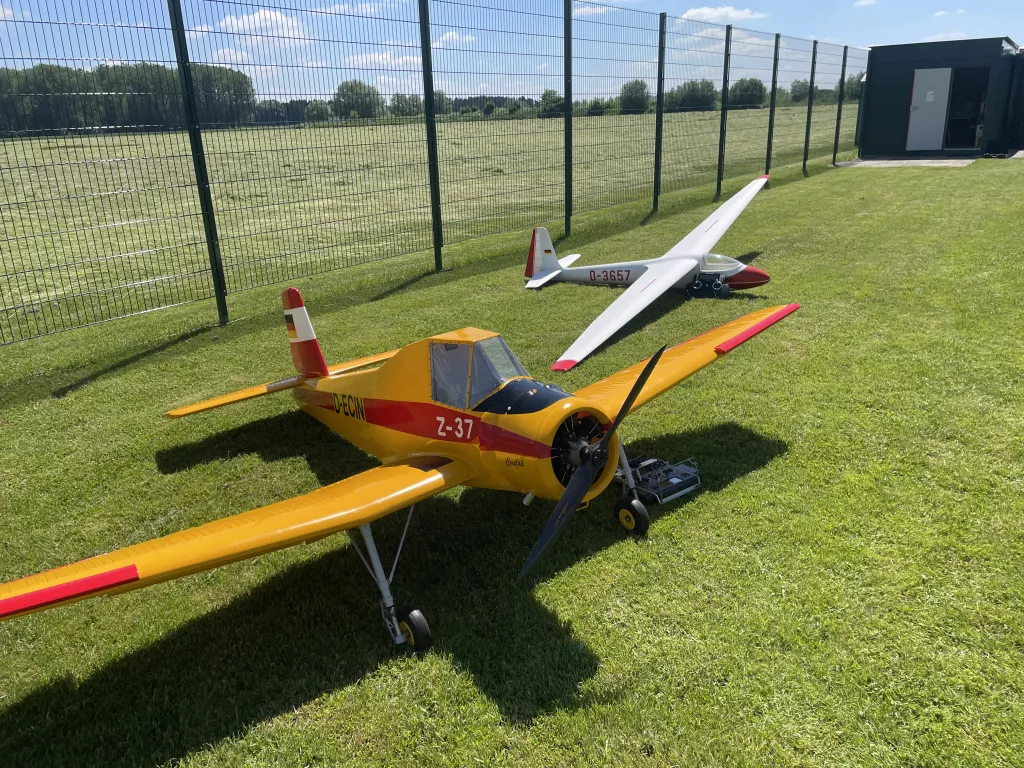 Gelbes Motorflugmodell und wei&szlig;es Segelflugmodell auf gr&uuml;ner Rasenfl&auml;che, umz&auml;unter Vereinsflugplatz mit Geb&auml;ude im Hintergrund