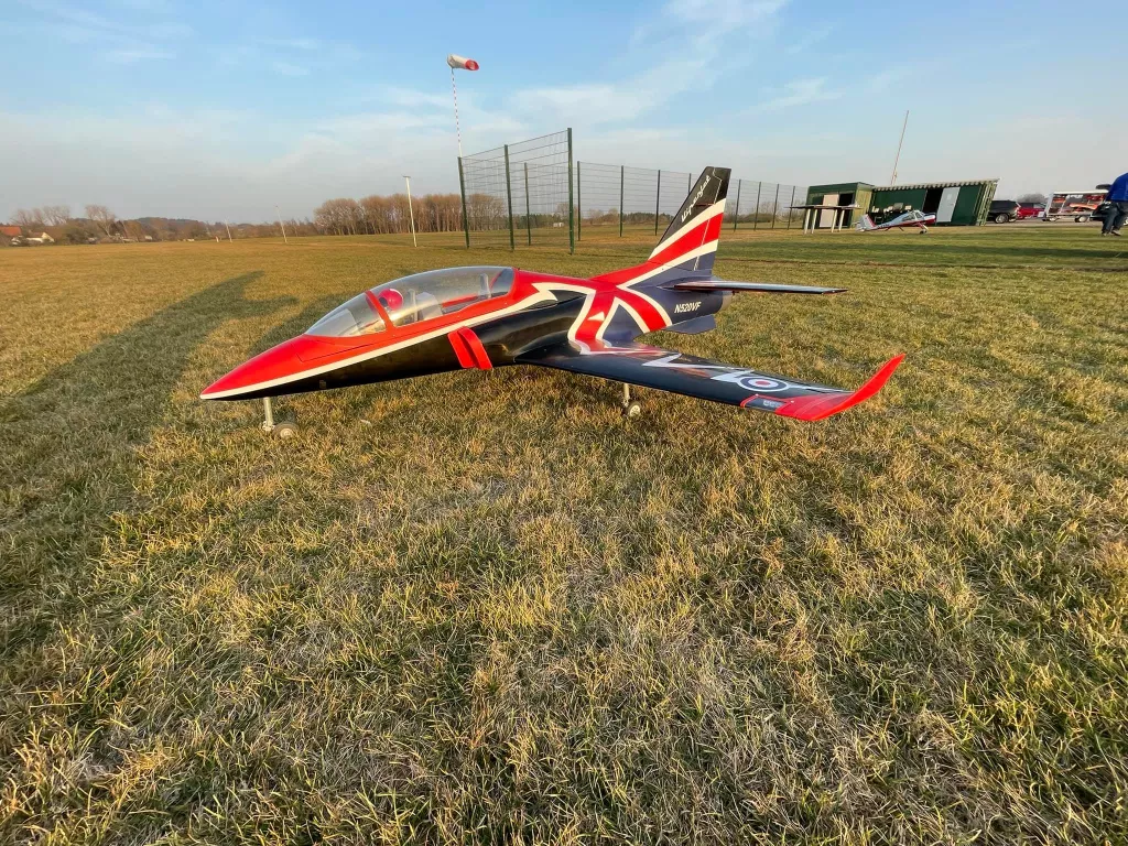 Rot-schwarzes Modellflugzeug auf Grasplatz, Windfahne im Hintergrund, Vereinsgeb&auml;ude sichtbar