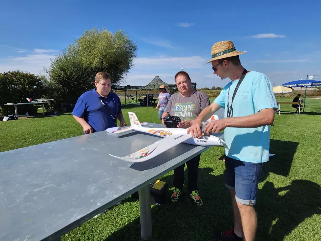Drei Personen an einem Tisch mit Modellflugzeug, Fernsteuerung, blauer Himmel, Rasenplatz im Hintergrund