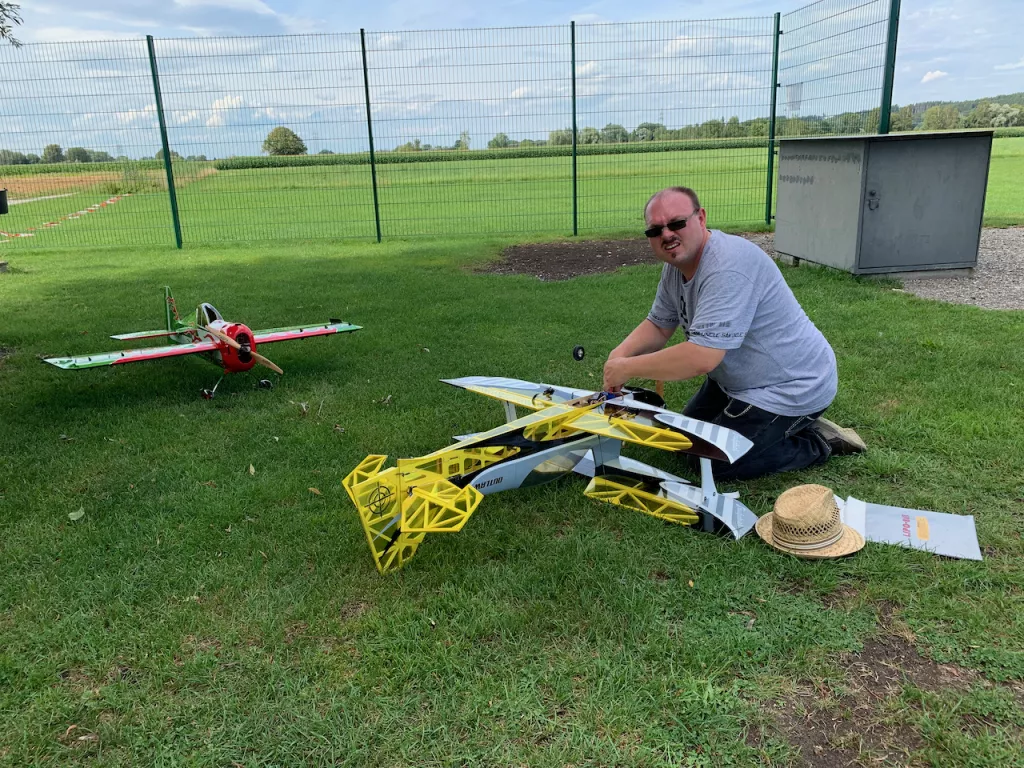 Zwei Modellflugzeuge auf gr&uuml;nem Rasenplatz, ein Mann in grauem T-Shirt sitzt daneben und arbeitet an einem gelb-wei&szlig;en Flugmodell