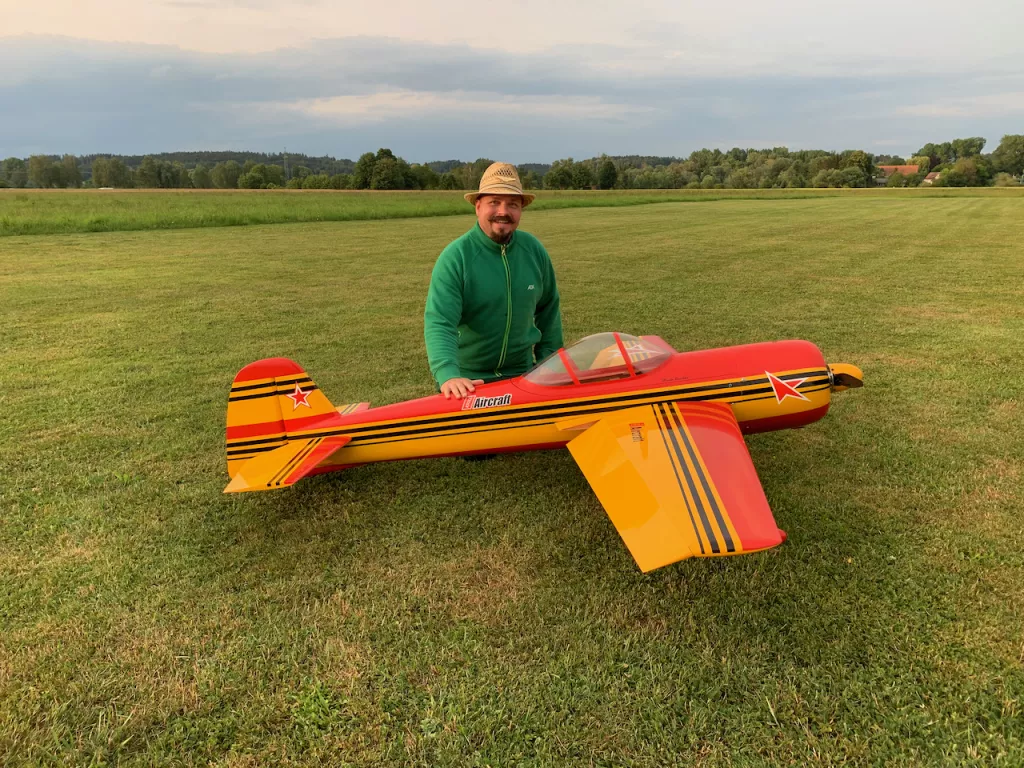 Rot-gelbes Modellflugzeug auf Rasenplatz, daneben l&auml;chelnder Mann mit Strohhut und gr&uuml;ner Jacke