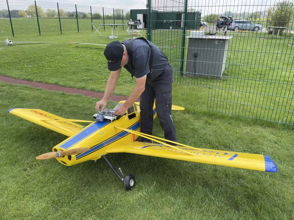 Modellflugzeug in gelb-blau auf Rasenfl&auml;che, Pilot justiert Fernsteuerung vor Vereinsgel&auml;nde