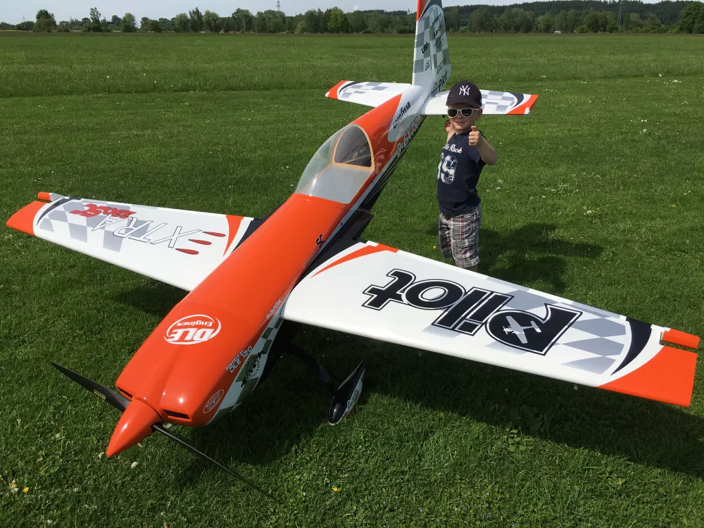 Orangewei&szlig;-gestreiftes Modellflugzeug auf gr&uuml;ner Wiese, daneben ein Kind mit Baseballkappe und Sonnenbrille, das Daumen hoch zeigt