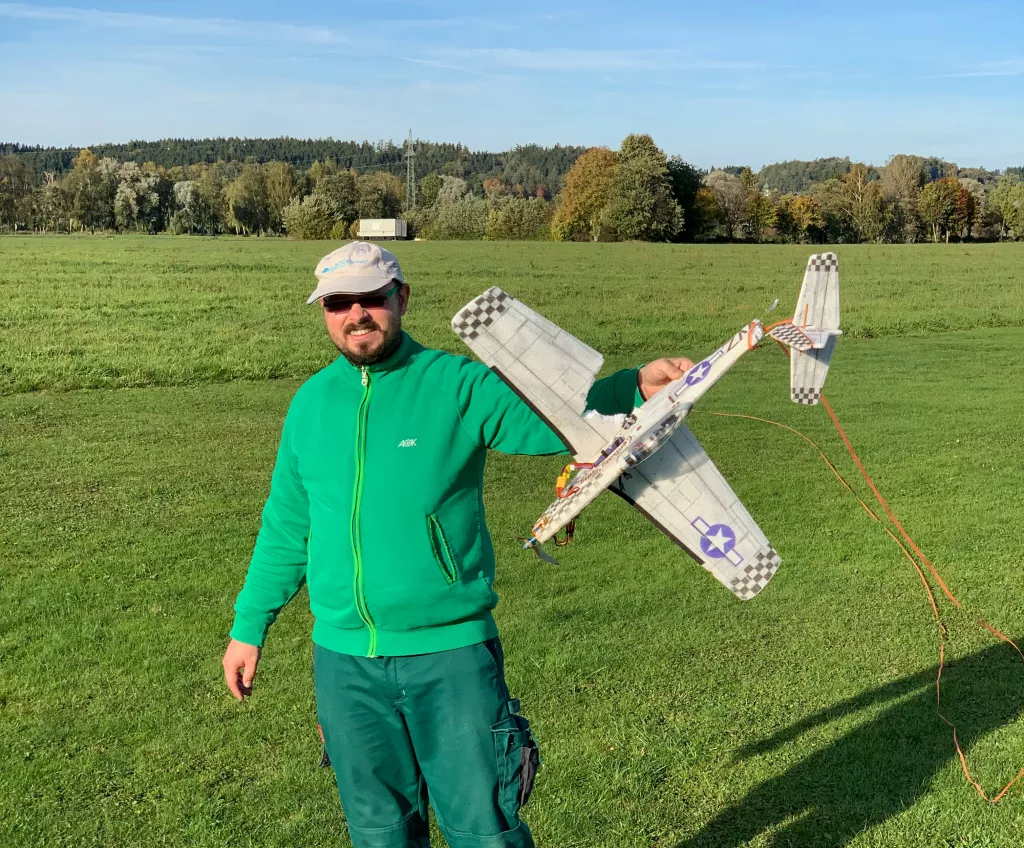Modellflugbegeisterter in grüner Jacke mit Flugzeugmodell auf Wiese, Herbstlandschaft mit Bäumen im Hintergrund
