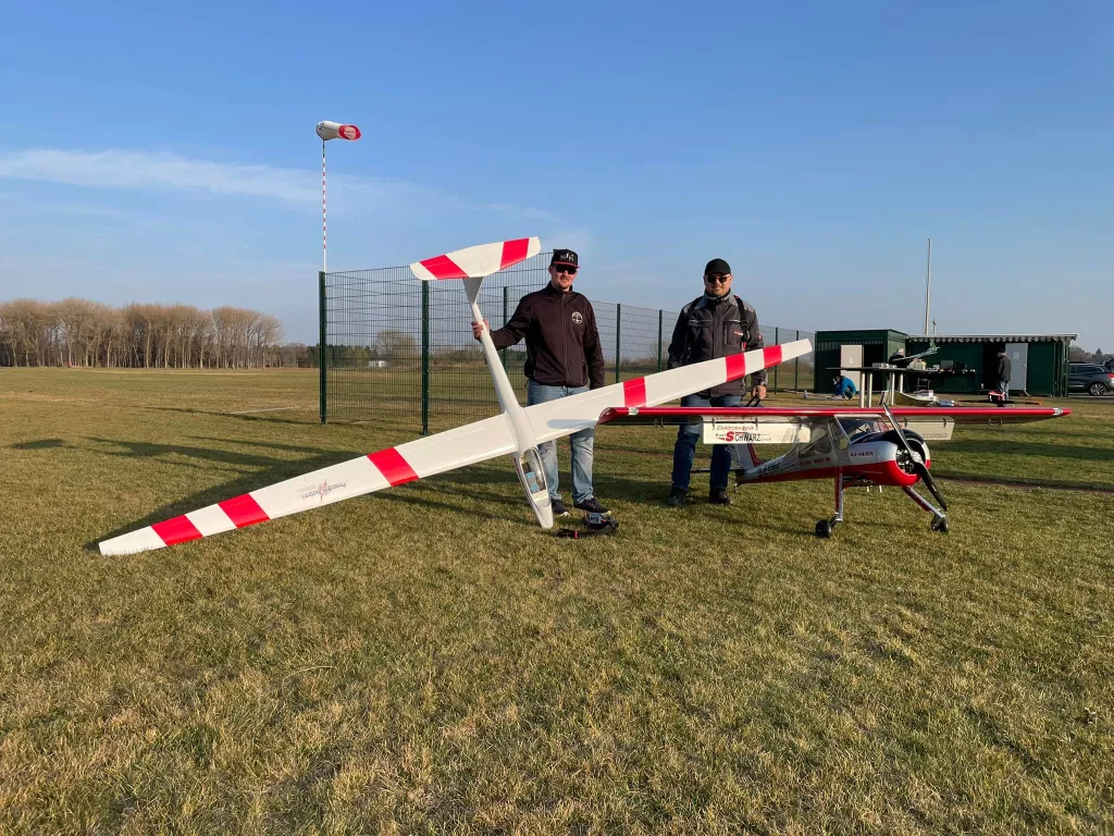 Zwei Modellflieger stehen neben einem gro&szlig;en wei&szlig;en Segelflugmodell mit rot-wei&szlig;en Fl&uuml;geln auf einer Wiese, im Hintergrund ein Flugplatz