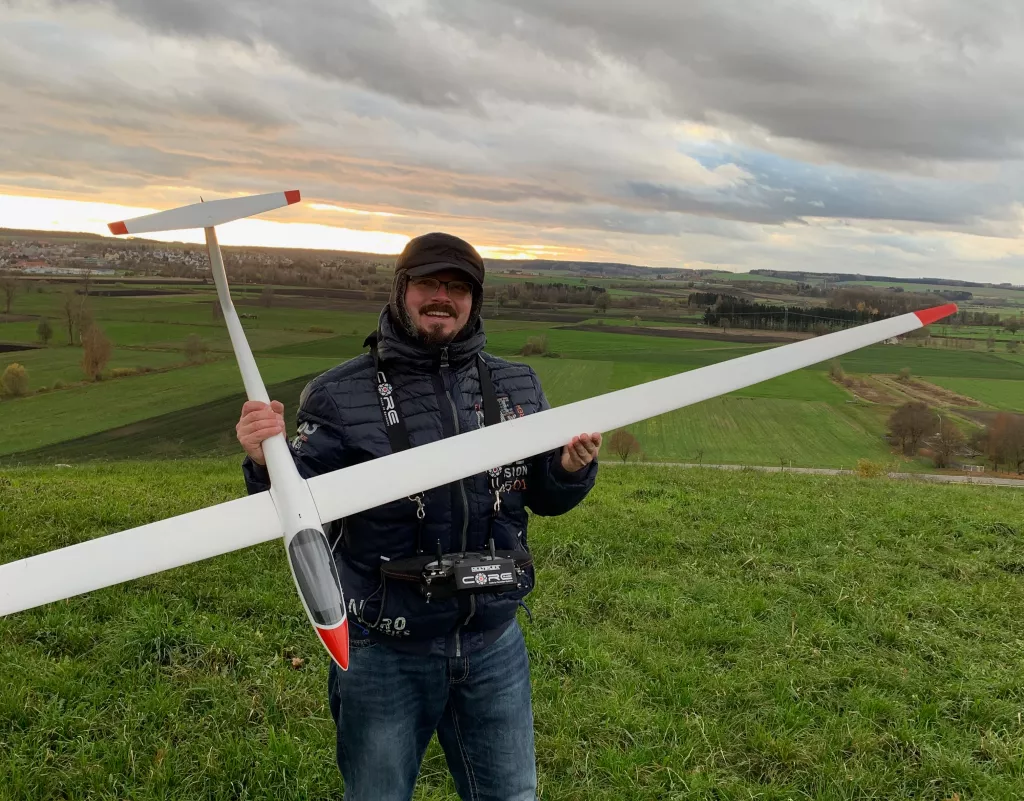 Person in Winterjacke h&auml;lt gro&szlig;es wei&szlig;es Segelflugmodell auf gr&uuml;ner Wiese, Landschaft mit Sonnenuntergang im Hintergrund