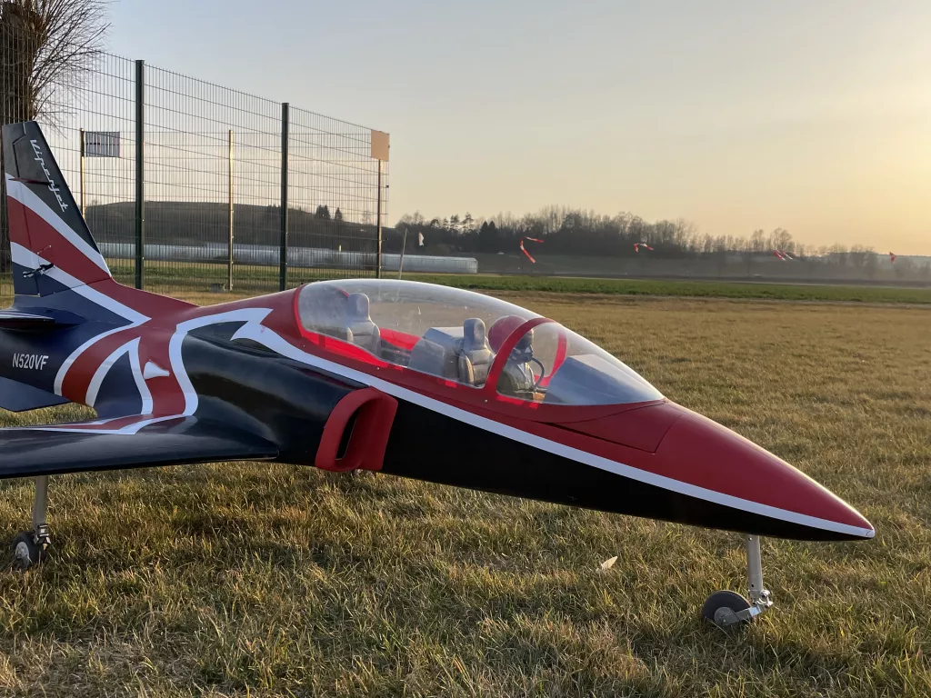 Modellflugzeug mit britischer Flaggen-Farbgestaltung, schwarz-rot-wei&szlig;, steht auf Graslandebene mit Zaun im Hintergrund