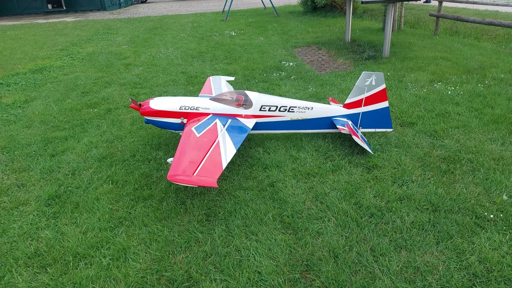 Rot-blau-wei&szlig;es Modellflugzeug vom Typ Edge 540 V3 steht auf gr&uuml;ner Rasenfl&auml;che mit Spielger&uuml;st im Hintergrund