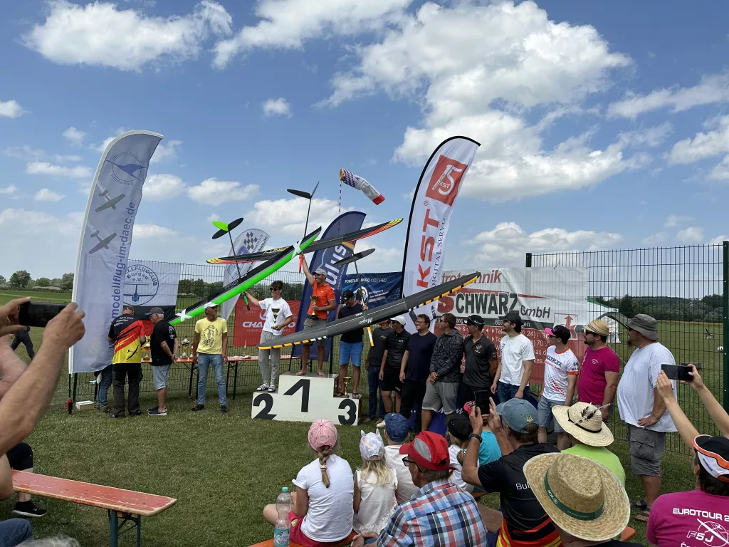 Podium bei Modellflug-Veranstaltung mit Siegern, Publikum und Vereinsfahnen vor blauem Himmel