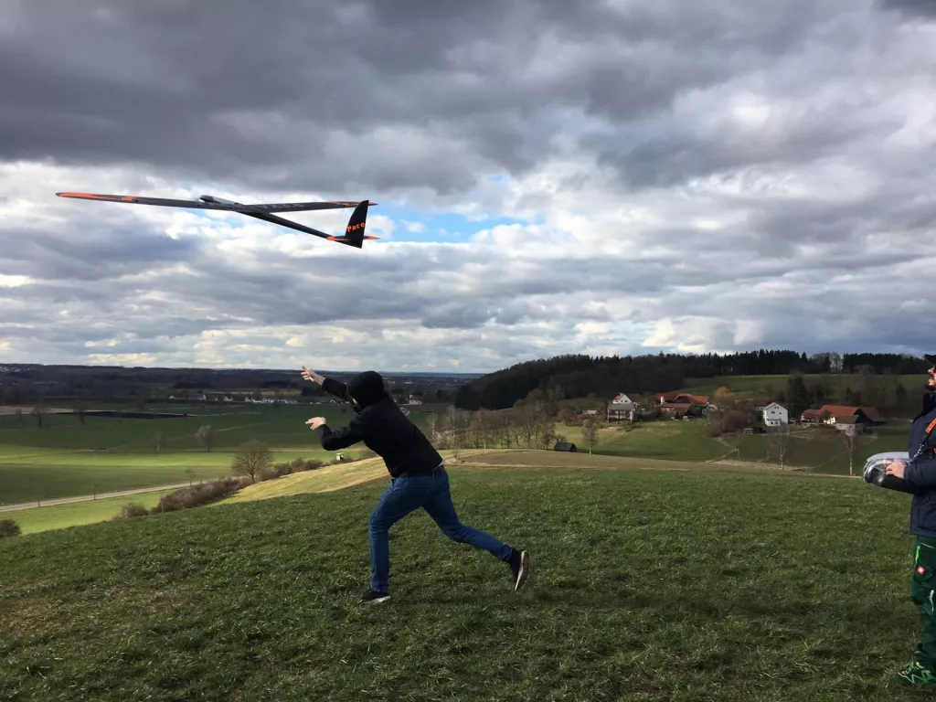 Modellflugzeug wird von einer Person auf einer grünen Wiese mit Häusern und Wald im Hintergrund gestartet