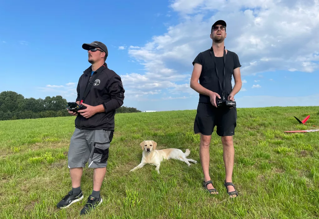 Zwei M&auml;nner mit Fernsteuerungen und gelbem Labrador auf gr&uuml;ner Wiese, blauer Himmel im Hintergrund