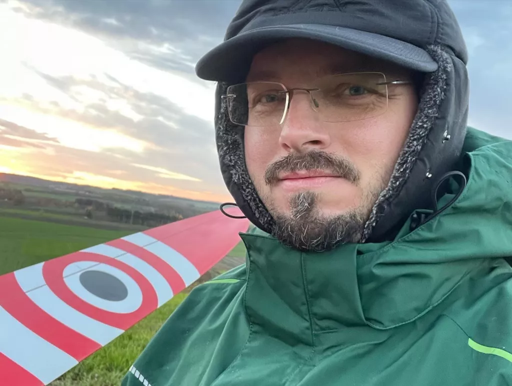 Person in gr&uuml;ner Winterjacke und M&uuml;tze bei Sonnenuntergang, im Hintergrund gr&uuml;ne Landschaft und rot-wei&szlig;e Flugzeugkante