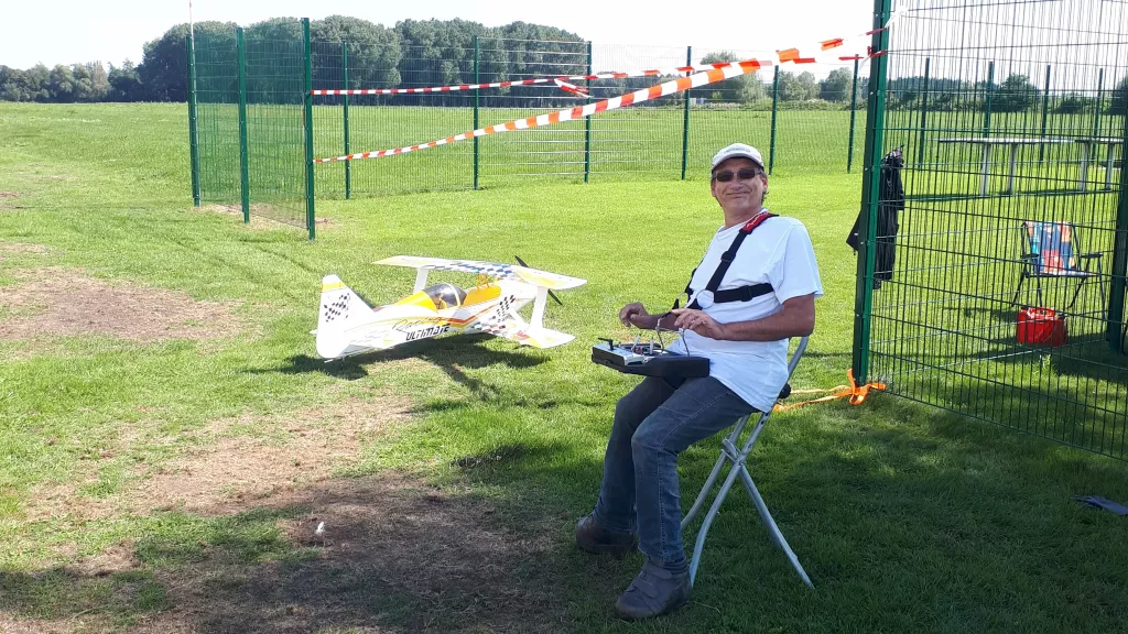 Modellflugpilot sitzt auf Klappstuhl mit Fernsteuerung, gelb-wei&szlig;es Flugzeugmodell auf Grasfl&auml;che, umz&auml;unter Vereinsplatz