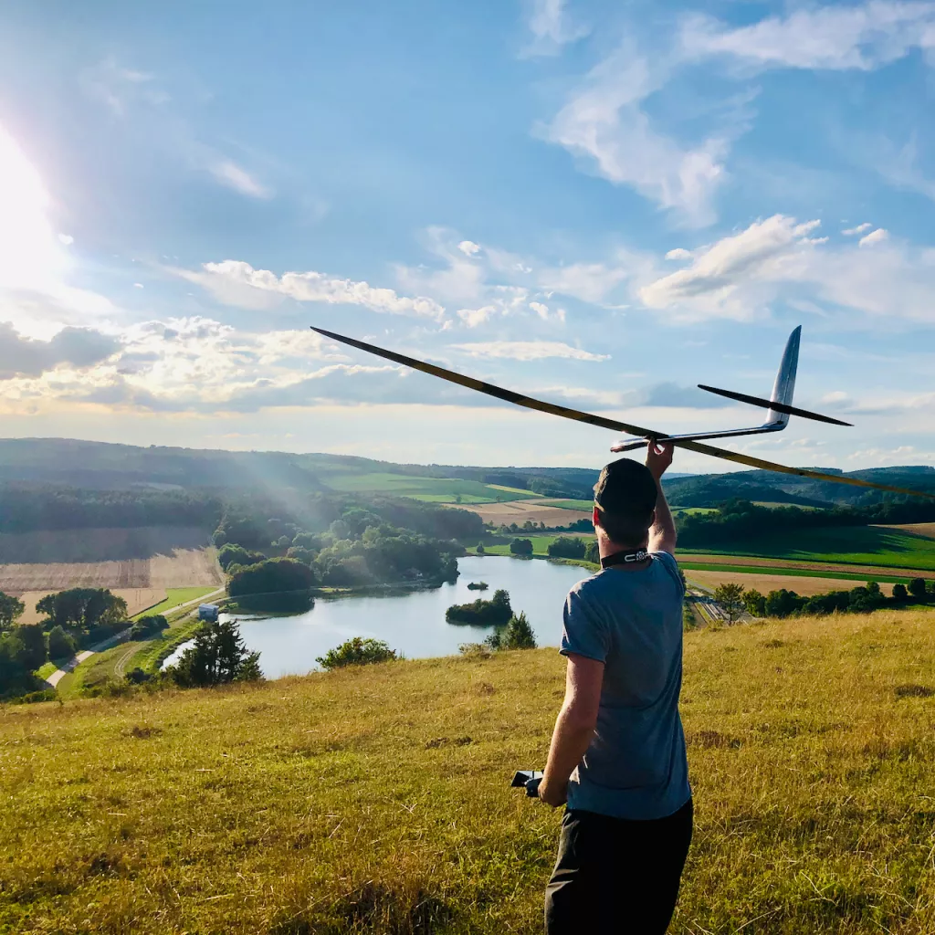 R&uuml;ckansicht einer Person mit Segelflugmodell &uuml;ber einer Wiese mit See und gr&uuml;ner H&uuml;gellandschaft im Hintergrund