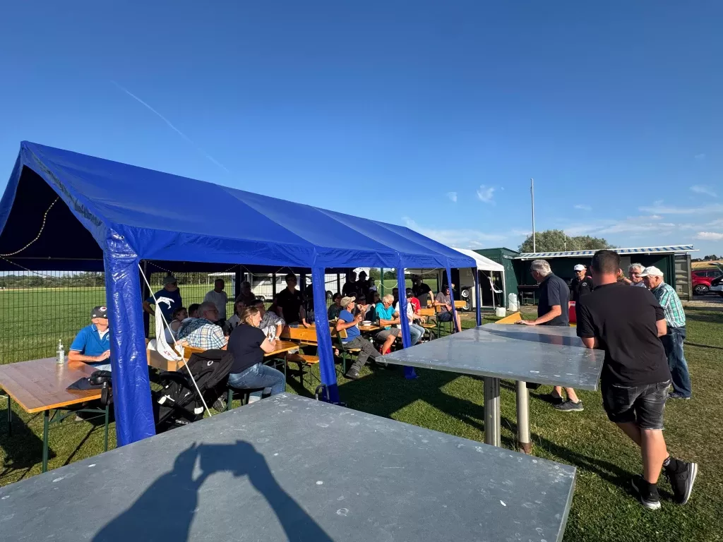 Blau &uuml;berdachter Pavillon mit Holztischen, mehrere Personen sitzen und stehen bei Vereinstreffen auf Rasenfl&auml;che