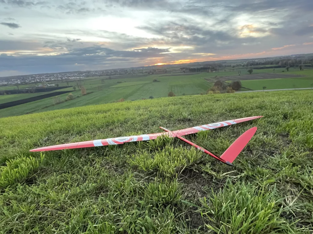 Rotes Segelflugmodell auf gr&uuml;ner Wiese, im Hintergrund Landschaft und dramatischer Abendhimmel mit Sonnenuntergang