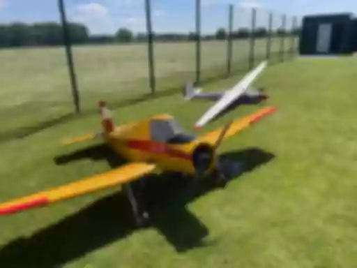 Gelbes Motorflugmodell und weißes Segelflugmodell auf grüner Rasenfläche, umzäunter Vereinsflugplatz mit Gebäude im Hintergrund