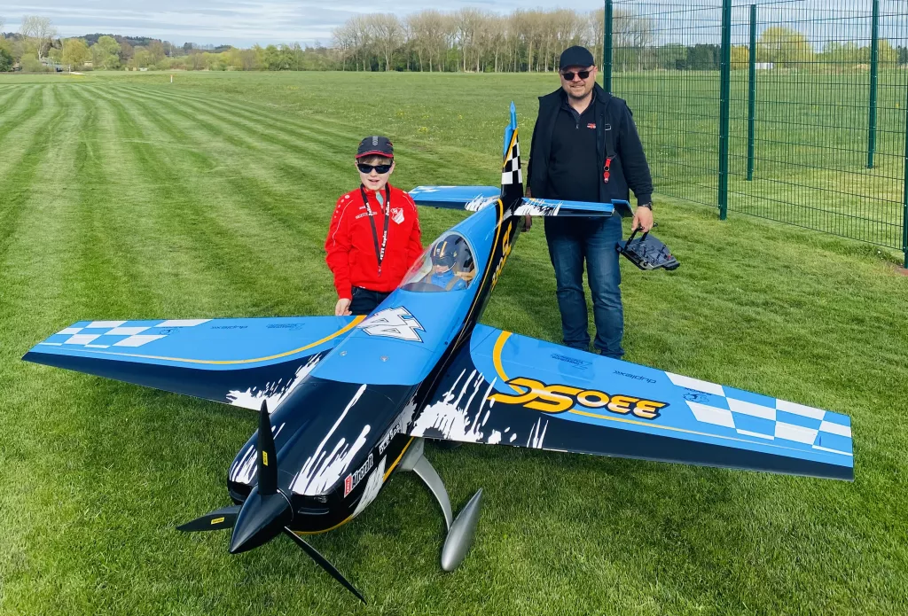 Blau-wei&szlig;es Modellflugzeug auf gr&uuml;ner Rasenfl&auml;che, daneben ein Kind in roter Jacke und ein Erwachsener