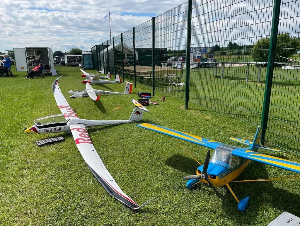 Mehrere Modellflugzeuge auf gr&uuml;ner Rasenfl&auml;che, darunter ein Red Bull Segelflugzeug und ein blau-gelbes Motorflugmodell, vor Maschendrahtzaun