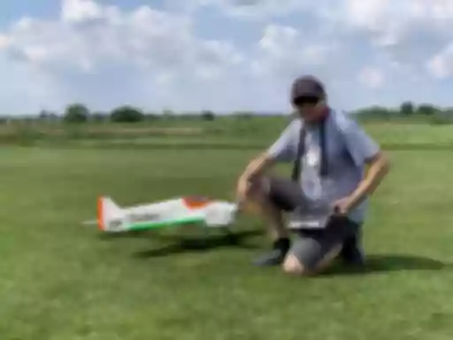 Modellflugzeug Rainbow in weiß-grün-orange, daneben kniender Modellflugpilot mit Fernsteuerung auf Rasenplatz