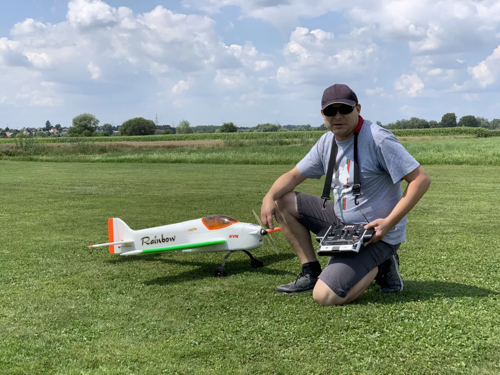 Modellflugzeug Rainbow in wei&szlig;-gr&uuml;n-orange, daneben kniender Modellflugpilot mit Fernsteuerung auf Rasenplatz