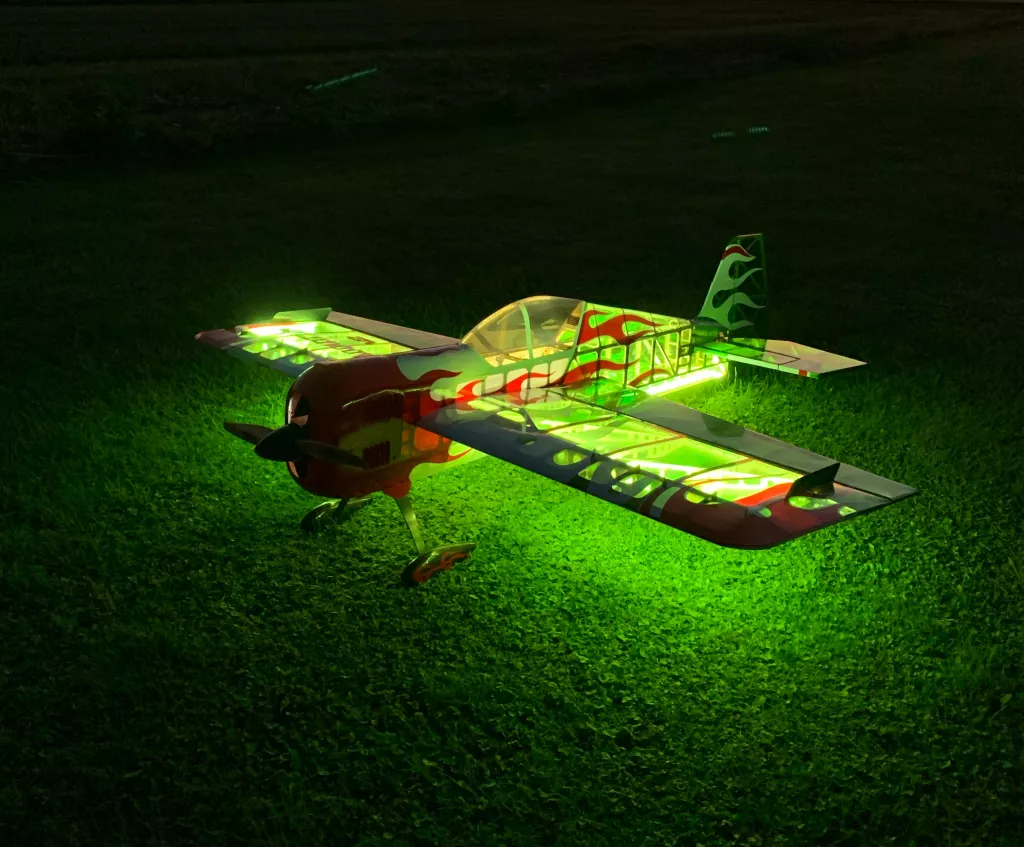 Beleuchtetes Modellflugzeug mit gr&uuml;n-roten Flammen-Design auf Grasnarbe, Kunstflug-Stil, Nachtaufnahme