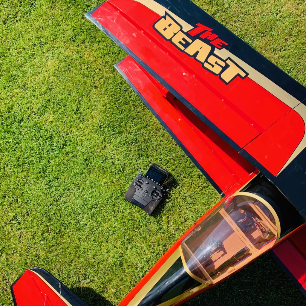 Rotes Segelflugmodell 'The Beast' mit schwarzen Fl&uuml;geln auf Rasen, daneben schwarze Fernbedienung