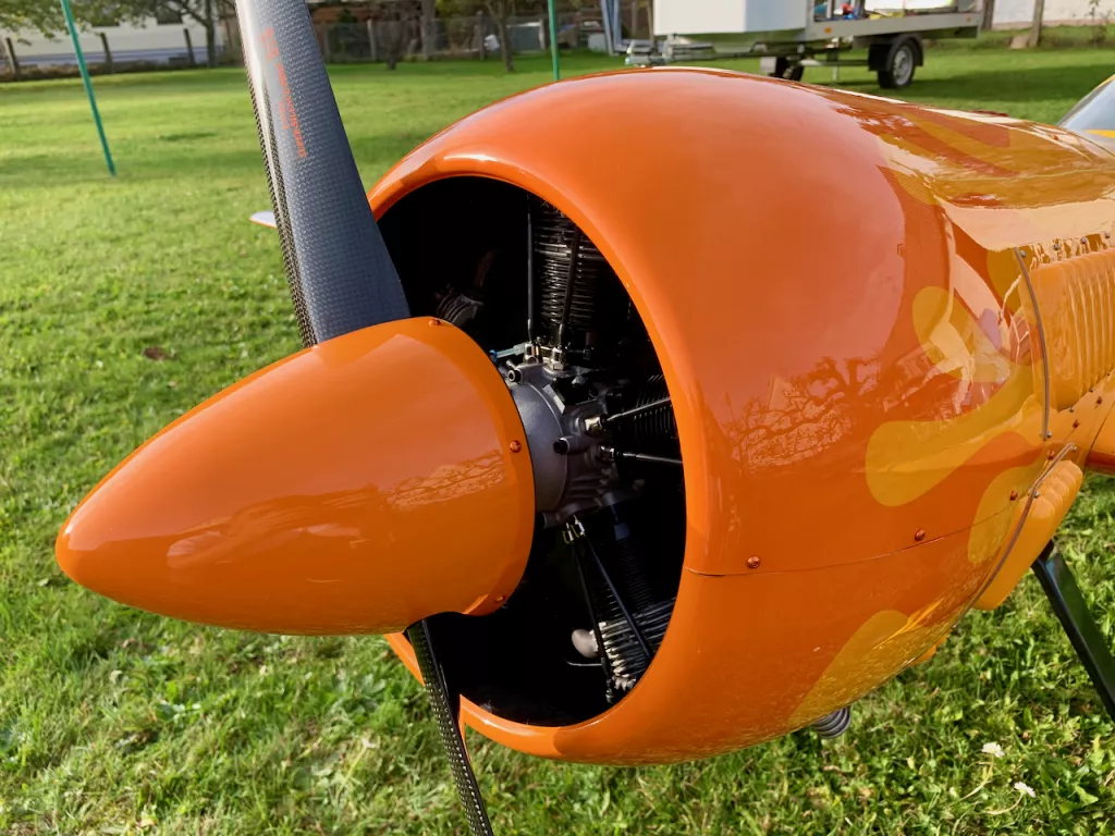 Nahaufnahme eines gl&auml;nzend orangefarbenen Flugzeugmotors mit Propeller, eingebaut in einen Modellflugzeug-Rumpf auf einer Wiese