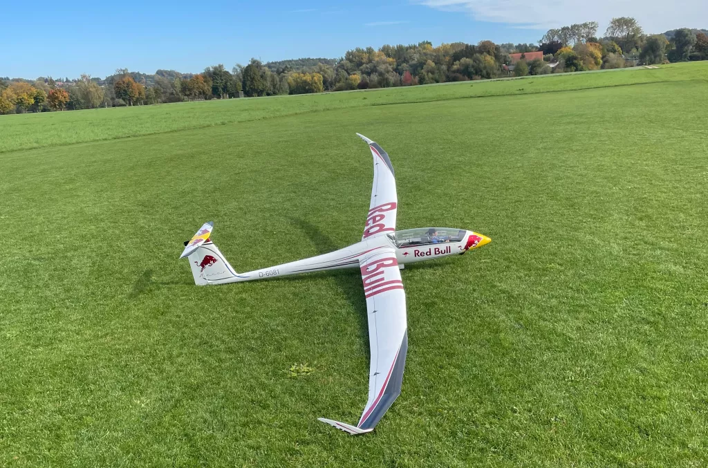 Wei&szlig;es Red Bull Segelflugzeugmodell mit ausgebreiteten Fl&uuml;geln auf gr&uuml;ner Wiese, Herbstlandschaft im Hintergrund