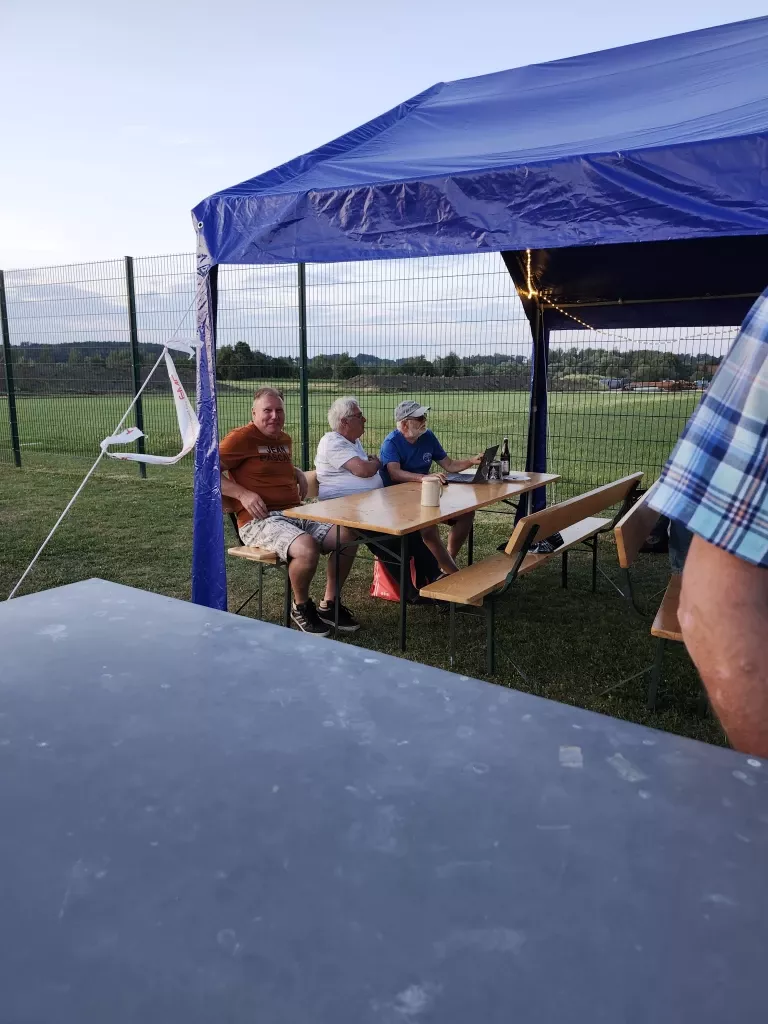 Drei Personen sitzen an Biertischen unter einem blauen Zelt auf einer Wiese mit Zaun und Landschaft im Hintergrund