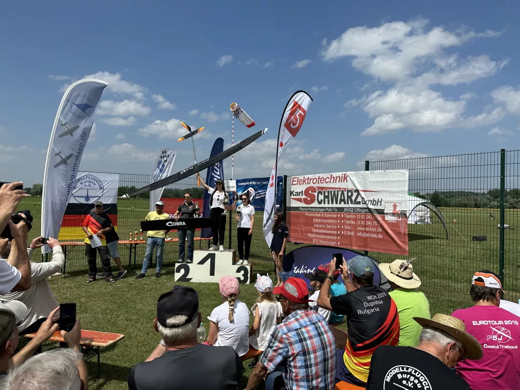 Siegerpodest bei Modellflug-Event, drei Personen auf Treppe, Zuschauer, Vereinsfahnen, Sommerhimmel