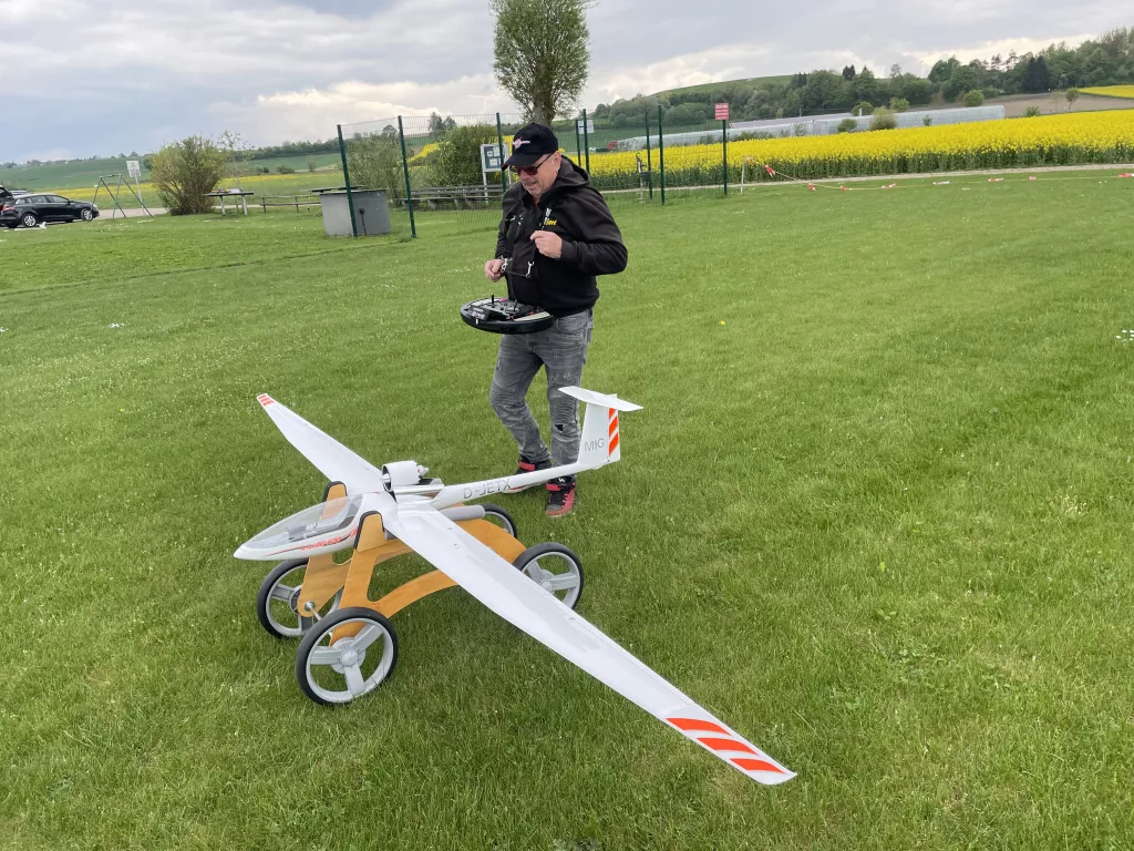 Gro&szlig;es wei&szlig;es Segelflugmodell mit gelb-orangen Elementen auf R&auml;dern, daneben ein Modellpilot mit Fernsteuerung auf Wiese