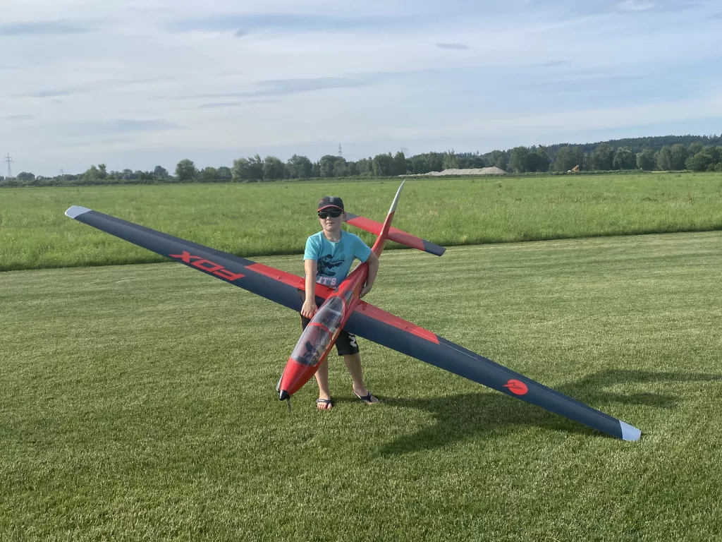 Rot-schwarzes Segelflugmodell auf gr&uuml;ner Rasenfl&auml;che, daneben stehend eine Person mit Sonnenbrille und Baseballcap
