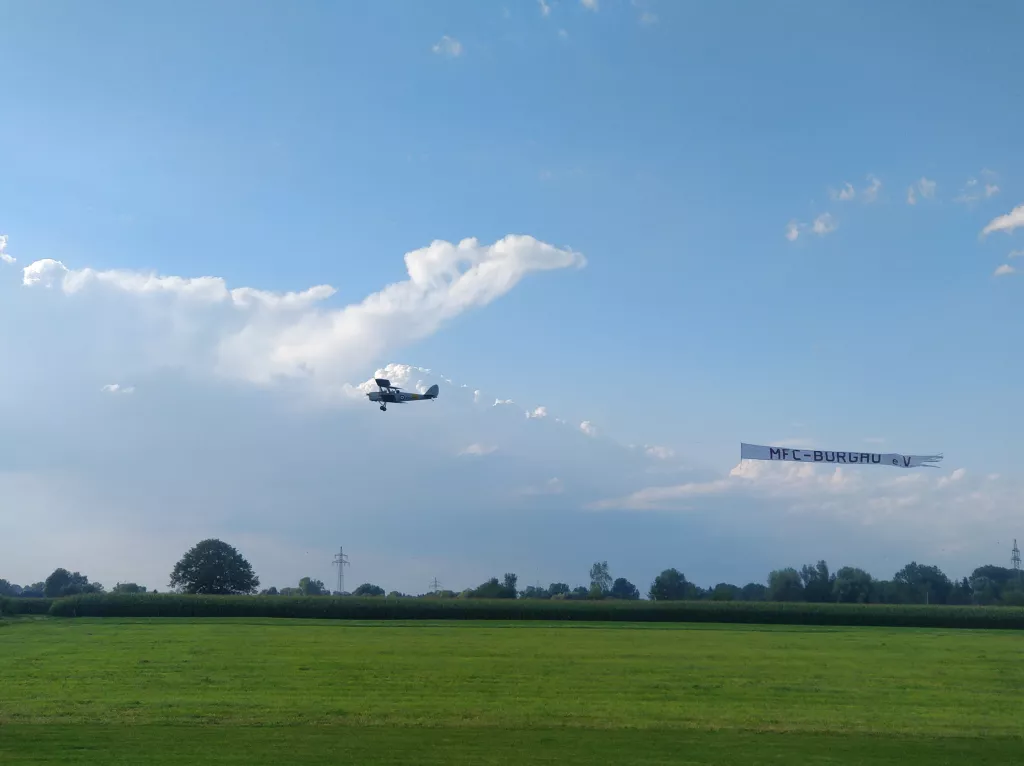 Kleines Modellflugzeug fliegt am blauen Himmel &uuml;ber gr&uuml;ne Wiese, Banner des MFC-Burgau e.V. rechts im Bild