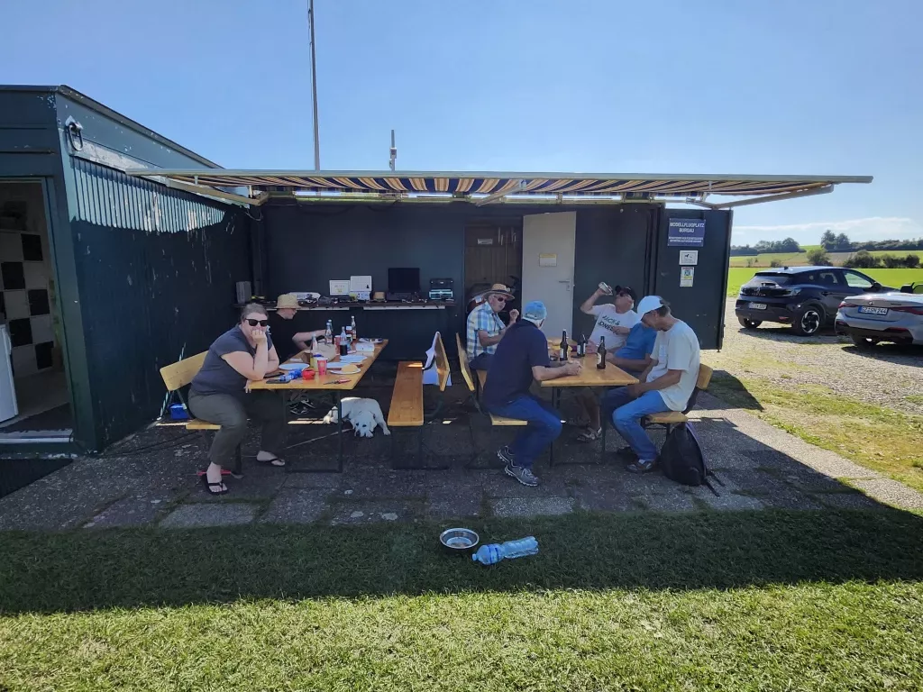 Mehrere Personen sitzen an Holztischen unter einem Vordach, Gr&uuml;nfl&auml;che, Autos im Hintergrund, Hund liegt am Boden