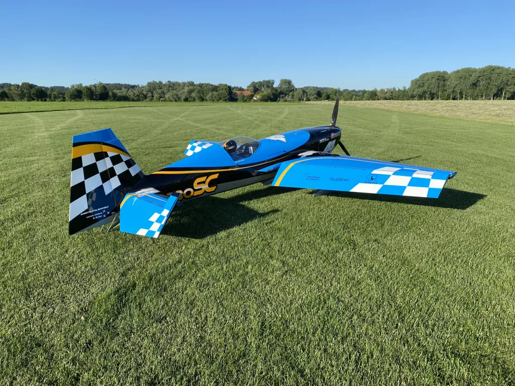 Hochdetailliertes Modellflugzeug in blau, wei&szlig; und schwarz mit Karomuster, geparkt auf sattgr&uuml;ner Wiese mit Baumkulisse
