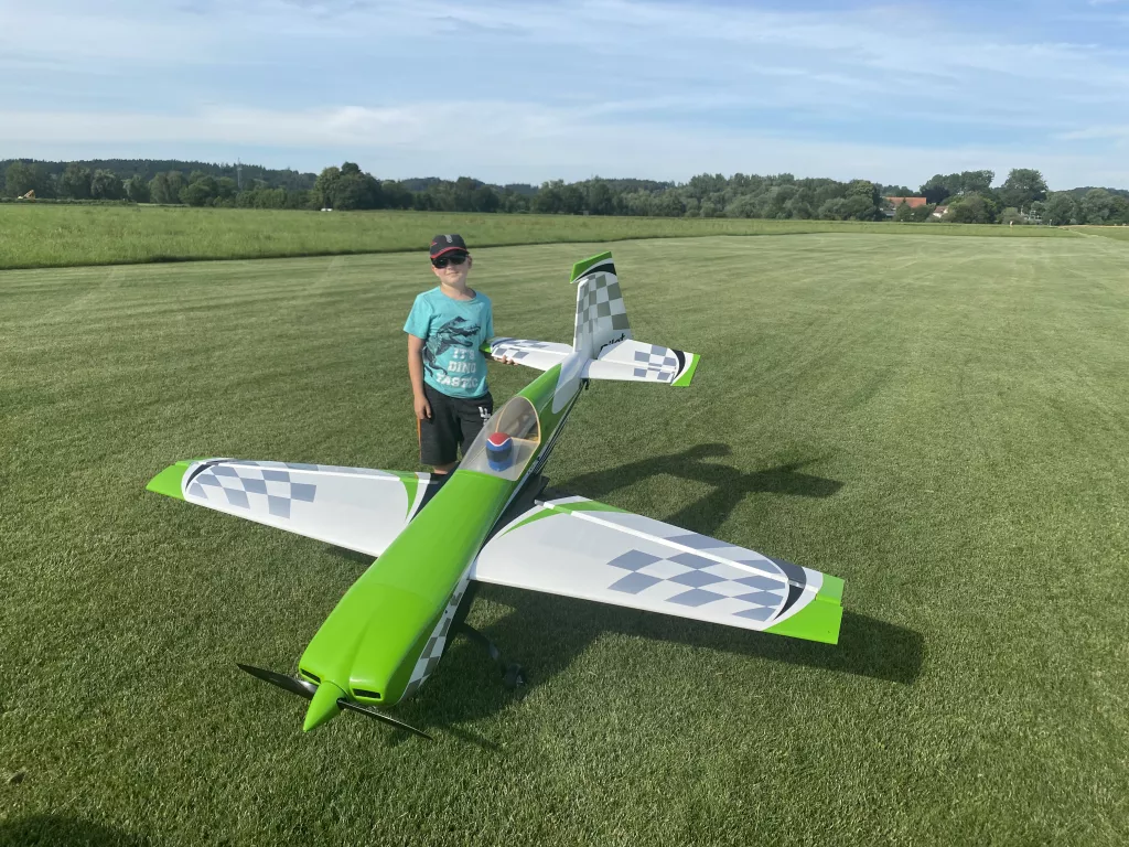 Gr&uuml;n-wei&szlig;es Modellflugzeug auf gem&auml;hter Rasenfl&auml;che, daneben eine Person in t&uuml;rkisem T-Shirt und Kappe