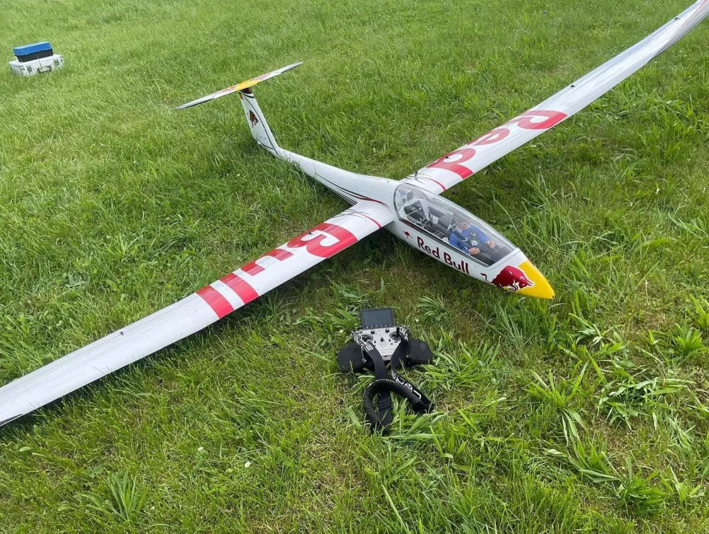Wei&szlig;es Segelflugmodell mit rot-wei&szlig;er Beklebung der Marke Red Bull, auf gr&uuml;ner Wiese liegend, mit Fernsteuerung im Vordergrund