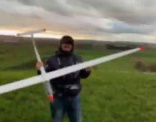 Person in Winterjacke hält großes weißes Segelflugmodell auf grüner Wiese, Landschaft mit Sonnenuntergang im Hintergrund
