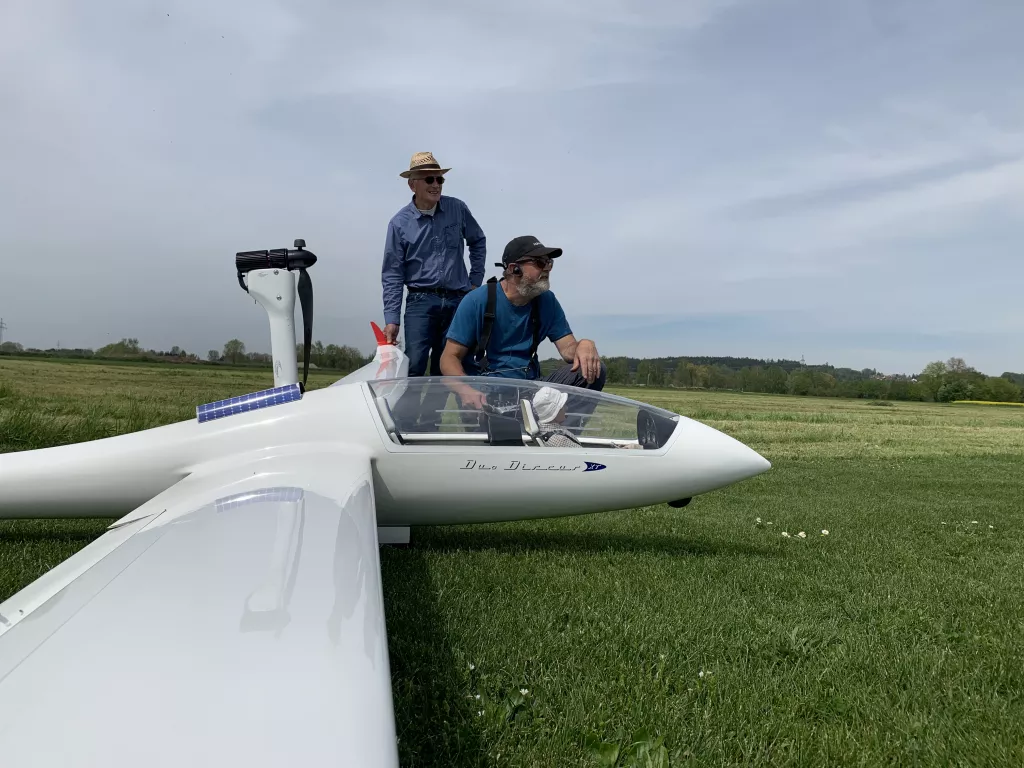 Wei&szlig;es Segelflugmodell Duo Discus auf Grasland, zwei Personen in Arbeitskleidung, eine Solar-Ladestation, blauer Himmel