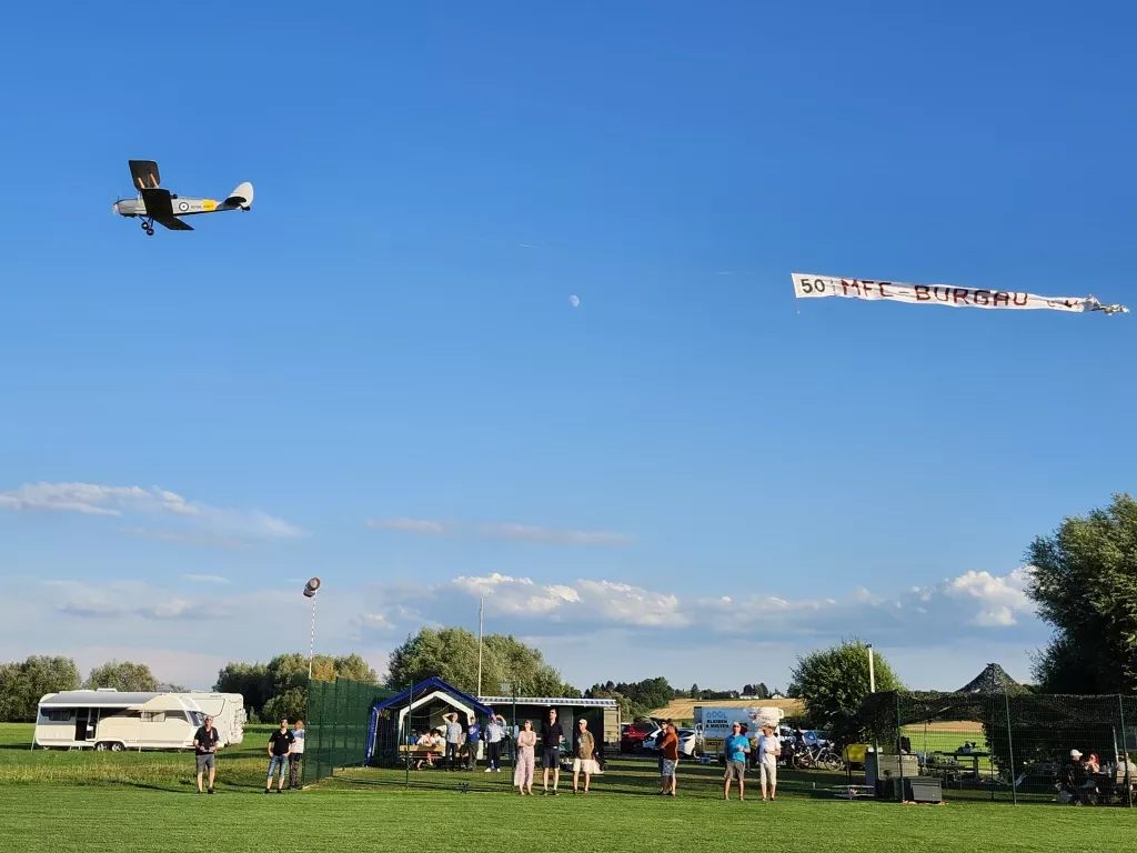 Historisches Modellflugzeug in der Luft, Transparent '50 MFC-Burgau', Vereinsmitglieder auf Grünfläche, Wohnwagen und Autos im Hintergrund